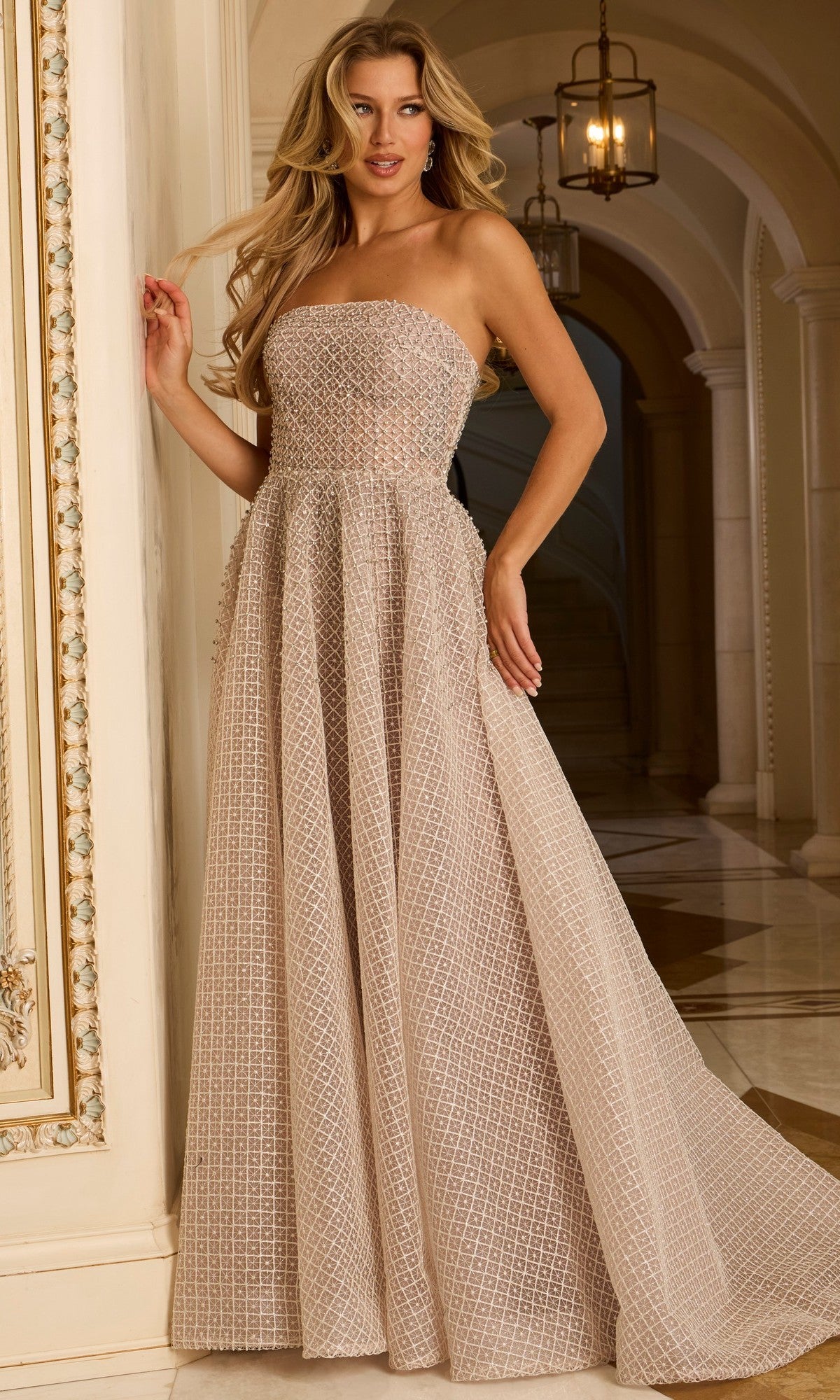 A woman wears a Jovani 45788 Long Prom Dress in Cafe with beaded grid texture, standing indoors near a decorative wall, marble floors, and an arched hallway in the background.